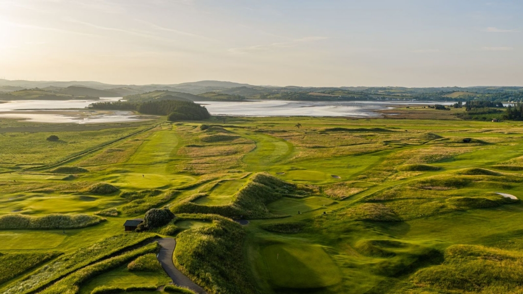 Golf course fairway and green with coastline and mountains in the background.