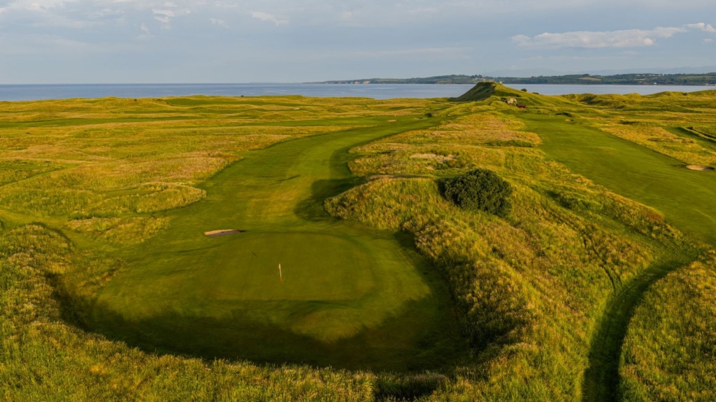 Golf course fairway and green surrounded by tall dunes, with rolling hills and mountains in the background